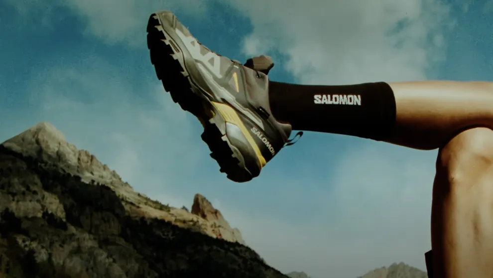 
            Ein Nahaufnahme-Foto eines Fußes in einem grauen und gelben Salomon-Trekkingschuh, der in der Luft schwebt. Im Hintergrund sind Berge und ein bewölkter Himmel zu sehen.