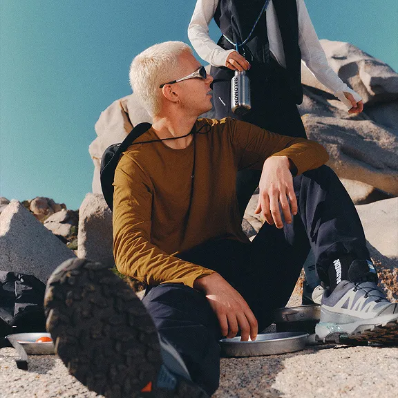 Wanderer in Outdoor-Kleidung sitzt entspannt auf Felsen, Salomon-Schuhe im Vordergrund.